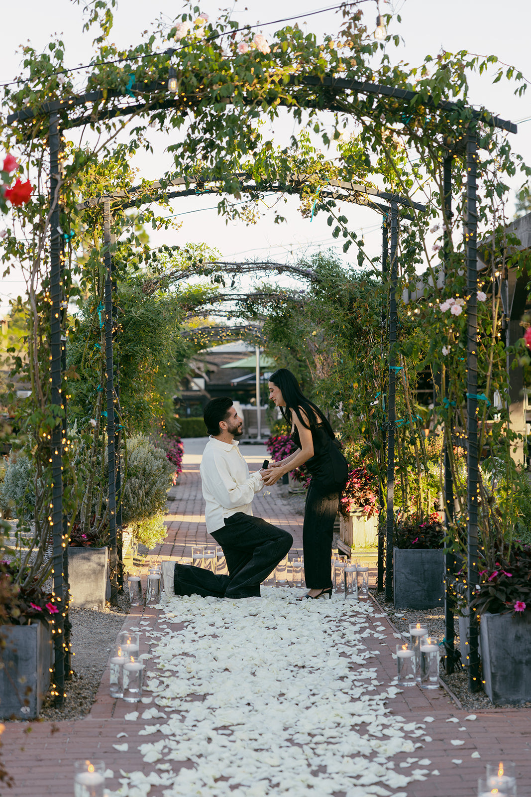Man kneeling to propose beneath vine-covered garden arches as his partner reacts